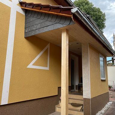 Gelbes Haus mit Windfang an welchem ein Vordach aus Holz nachträglich angebaut wurde und die Stirnseite mit Schiefer verkleidet wurde.