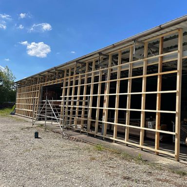 Holzlattung eines Gebäudes als Vorbereitung für die spätere Fassade unter klarem blauen Himmel. Baumaterialien sind sichtbar.
