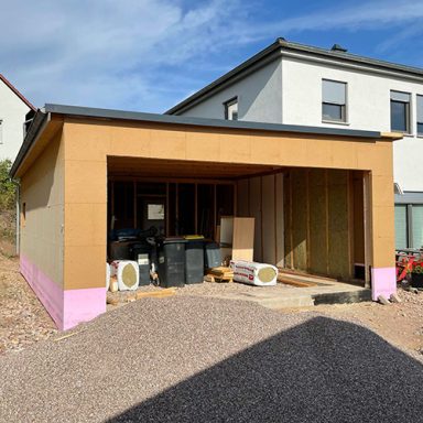 Ein neugebauter Garagen-Anbau mit Holzwänden und pinkem Sockel, umgeben von Schotter.