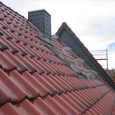 Dach mit roten Ziegeln und einem schieferverkleidetem Schornstein, teilweise bewölkter Himmel.