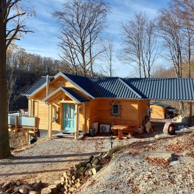 Massivholz-Hütte mit blauen Dach, umgeben von Bäumen und einem gepflasterten Weg.