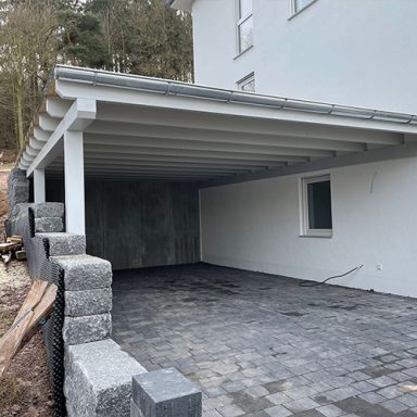 An ein Haus angebauter Doppelcarport mit grauen Steinplatten und einer weißen Wand im Hintergrund.